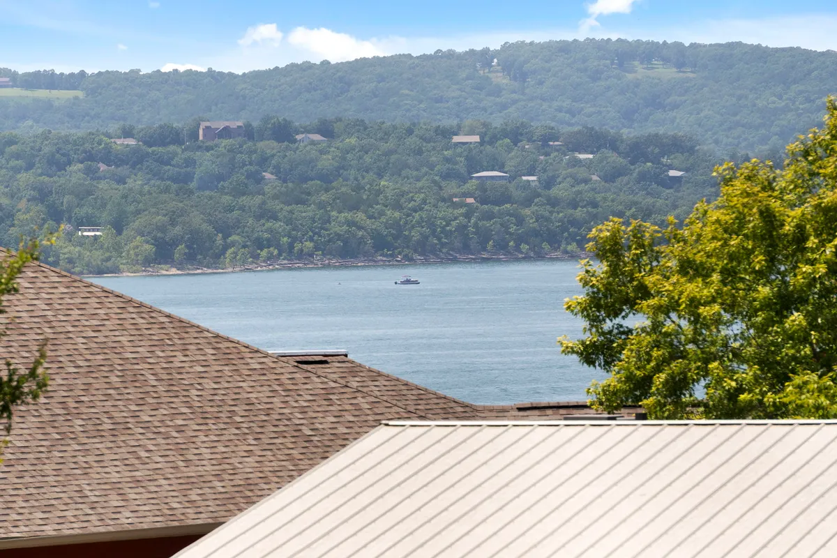 This image presents a peaceful view of the lake with mountains in the distance, rooftops of lakeside homes, and lush trees in the foreground. The scene captures the natural beauty and recreational possibilities of the lake and its immediate surroundings.