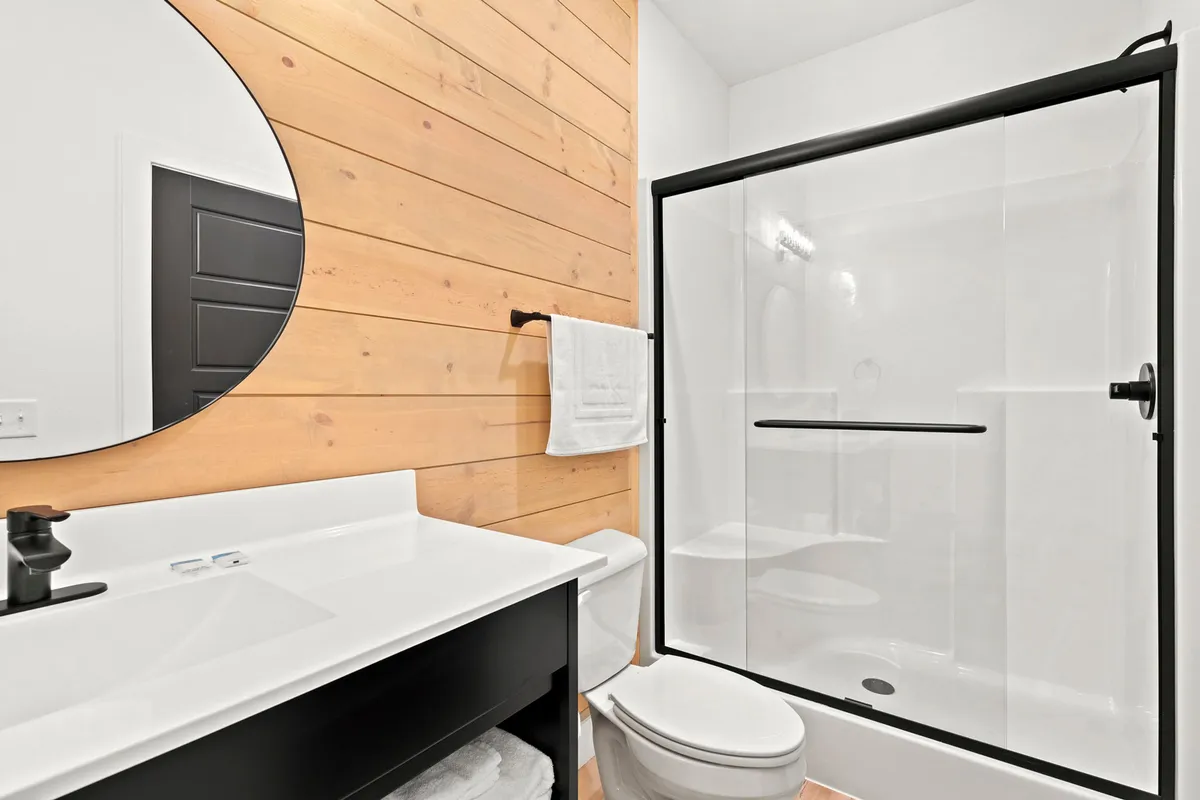A stylish, spa-inspired bathroom with a black-framed glass shower, round mirror above the white sink, and a wooden accent wall. The space feels calm and refreshing, echoing a high-end hotel experience.