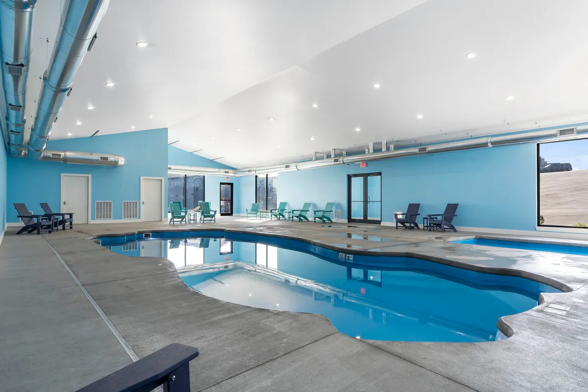 A bright, modern indoor swimming pool with refreshing blue walls and comfortable lounge chairs lining the poolside. The calm water reflects the light, creating an inviting atmosphere perfect for both relaxation and socializing.