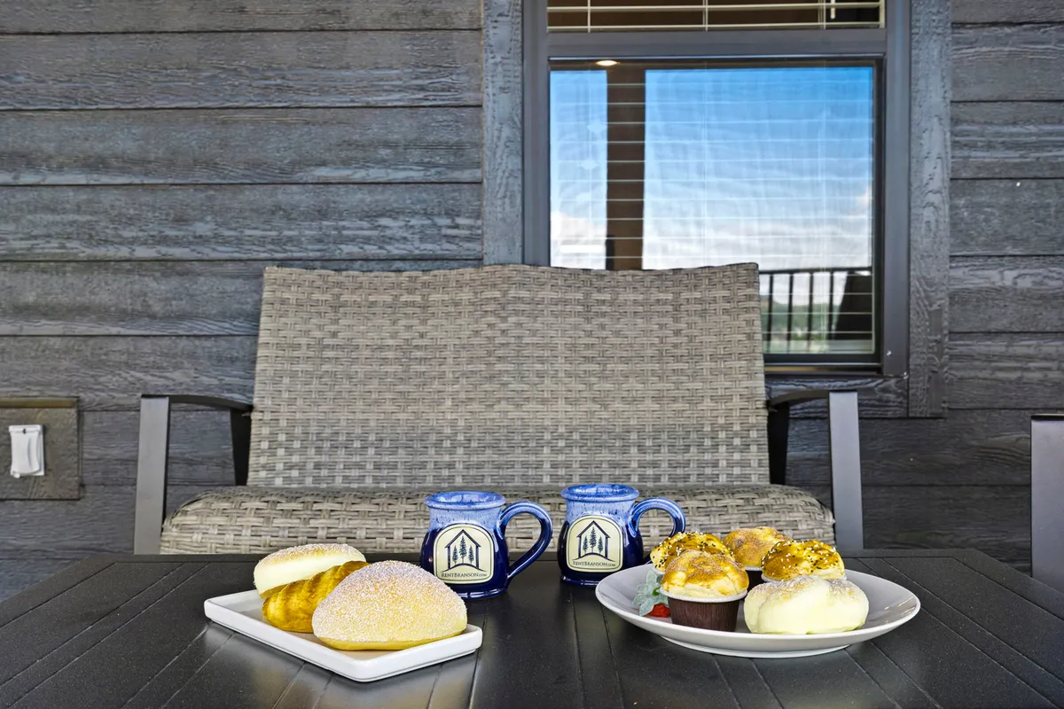 A cozy exterior porch scene featuring a woven chair and a breakfast spread with blue mugs, with reflections in the window hinting at the inviting interior beyond. The space is set for relaxation, socializing, and enjoying the morning sun, with easy access and a welcoming atmosphere.