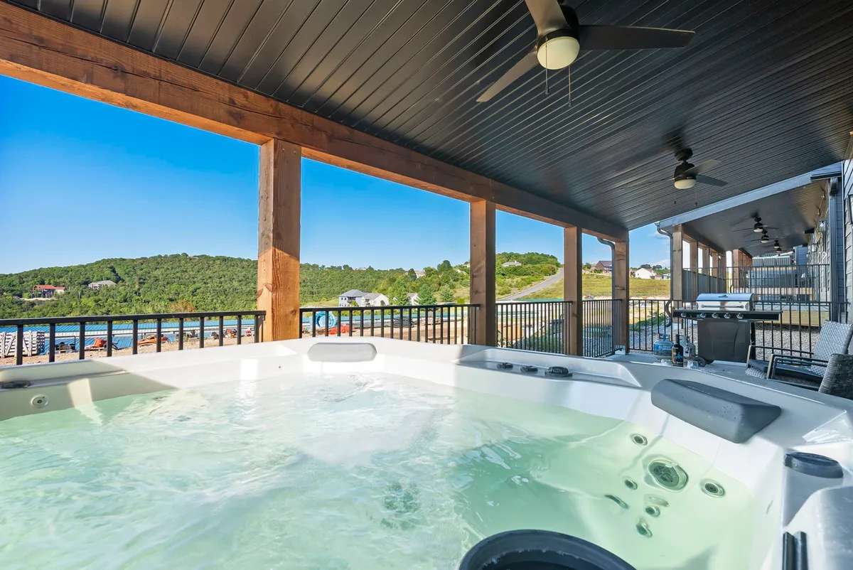 The image captures the exterior of a newly built single-family home at sunset, with elegant wooden beams framing a spacious deck. A bubbling hot tub sits invitingly in the foreground, overlooking sweeping lake vistas and rolling hills, evoking a peaceful, luxurious atmosphere. The serene mood is perfect for relaxing, entertaining, or stargazing under the open sky.
