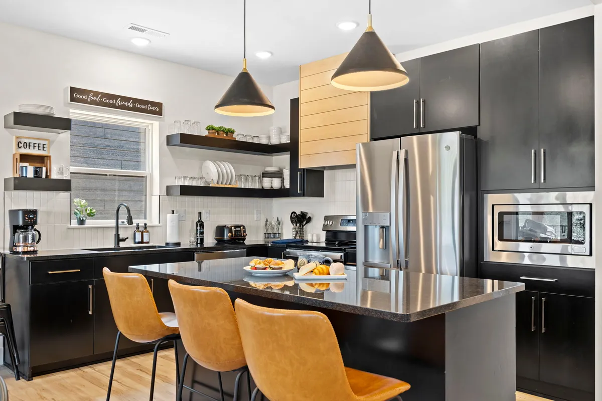 A modern kitchen with a central island and bar seating, black cabinetry, hanging pendant lights, granite countertops, and open shelving with neatly arranged dishes. The space is ideal for both cooking and socializing, encouraging shared culinary experiences.