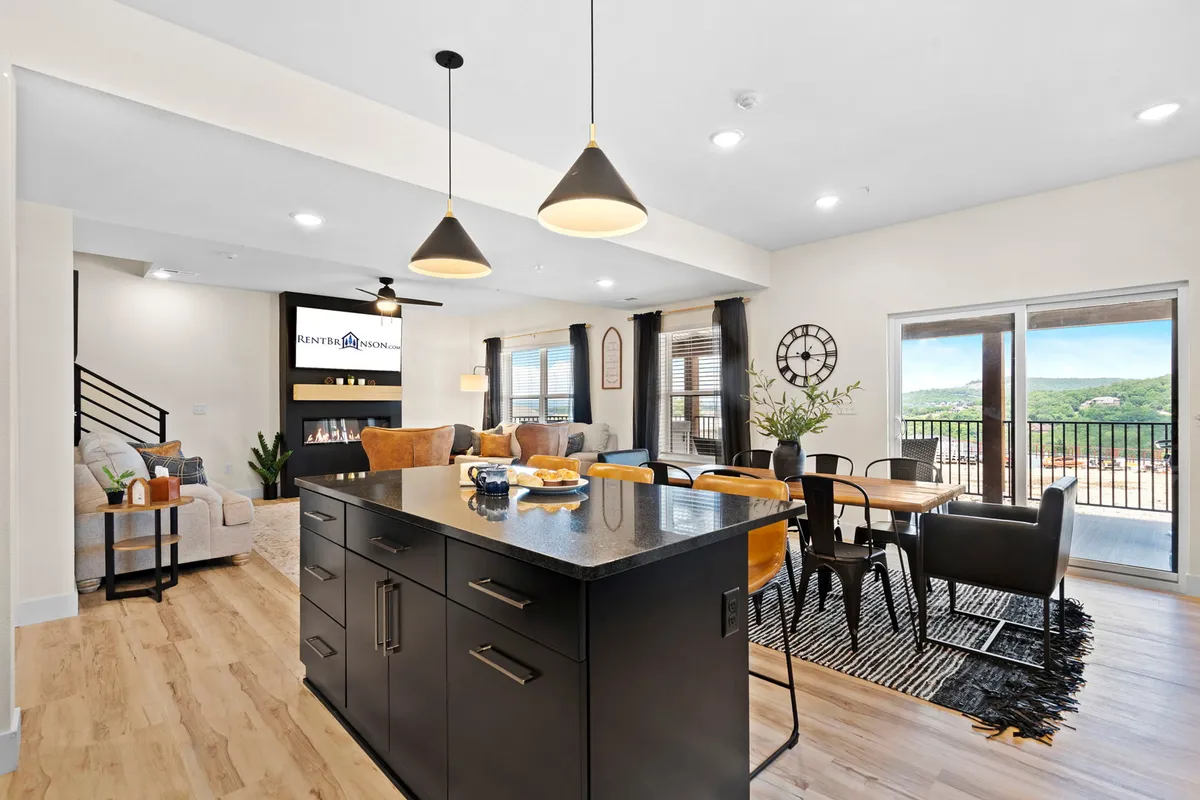 A wide, open-concept space that seamlessly combines the kitchen, dining area, and living area. The central kitchen island forms the hub, with views stretching to the dining table and a comfortable living area with plush seating. Large windows let in abundant natural light and offer scenic views of the outdoors, creating a welcoming atmosphere for gatherings.