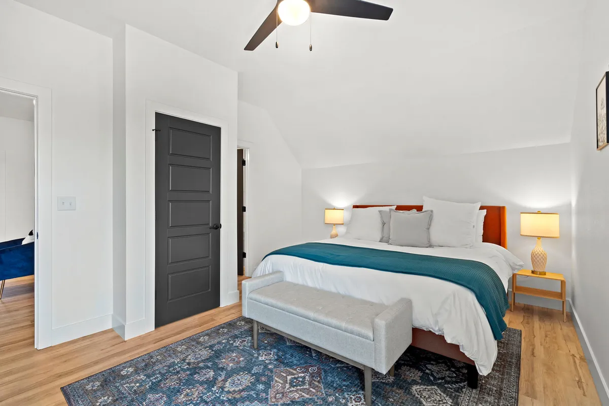 A serene king-sized bedroom featuring a large bed with decorative throw pillows, nightstands with lamps on either side, and a soft area rug beneath. Natural light fills the room, illuminating tranquil decor and modern touches.