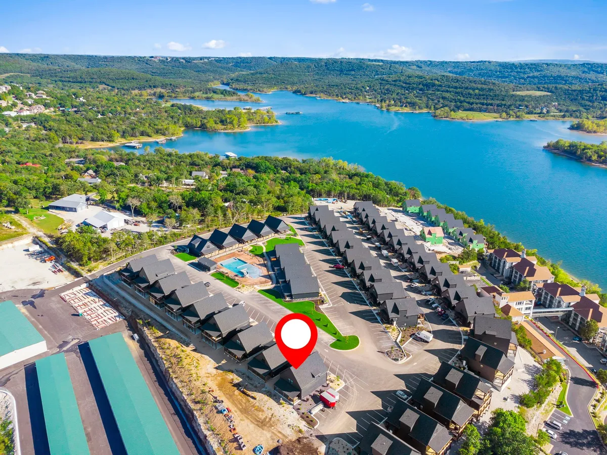 A scenic lakefront area featuring a swimming pool, residential buildings, landscaped lawns, and children playing. The backdrop includes Branson Bay Marina and boats on the water, hinting at easy access to marina activities.